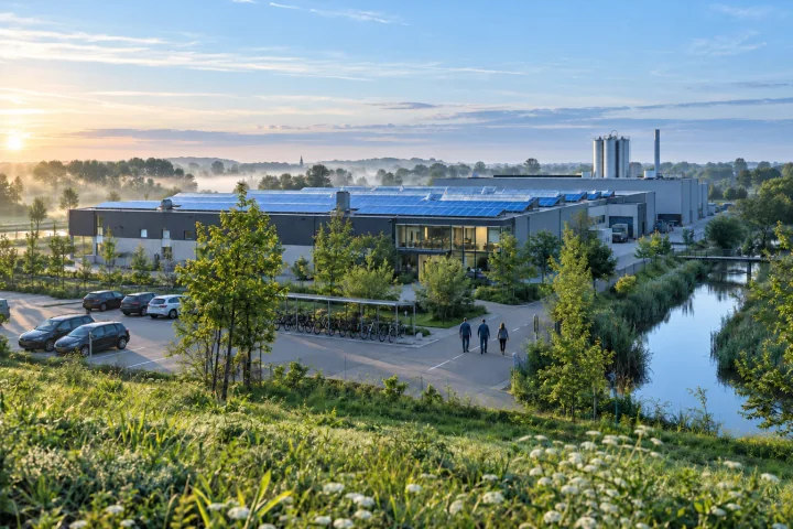 Three employees walking from the car park towards a factory with solar panels on its roofs, beside a canal and bicycle racks at misty dawn