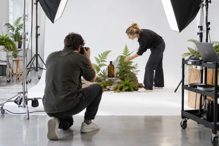 Ein Fotograf kniet, um eine Flasche aus Bernsteinglas zu fotografieren, während eine Stylistin in einem hellen Studio Farne und Moos darum herum anordnet