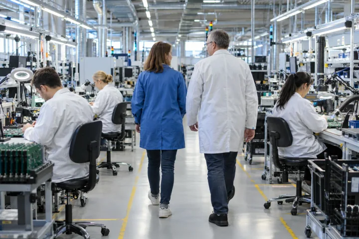 A visiting auditor in a blue coat and a supervisor walking between workbenches where workers assemble circuit boards