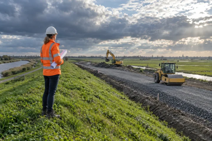 Une ingénieure civile avec des plans enroulés observe une pelleteuse et un compacteur à l’œuvre sur une digue néerlandaise